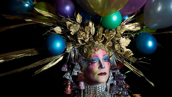 A headshot of a person with an elaborate head piece of golden flora and multicolored balloons, and brightly colored stage makeup